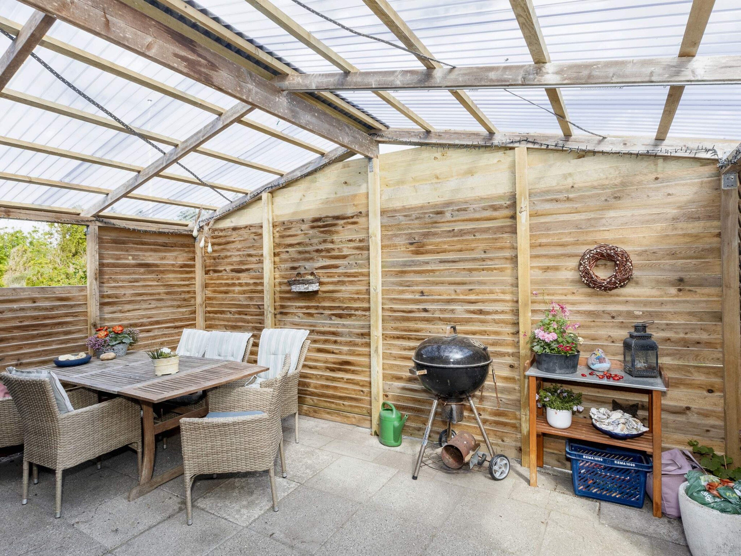 Un portico coperto con un tavolo e sedie di legno, un barbecue e un piccolo ripiano con piante e oggetti decorativi.