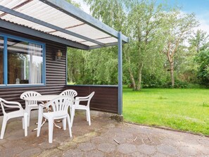 A wooden cabin with a covered patio, a table with chairs, and a view of a green landscape.