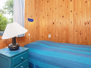 Una habitació amb paret de fusta, un llit amb mantell blau i blanc a ratlles, una tauleta de nit amb una làmpada i una presa elèctrica.