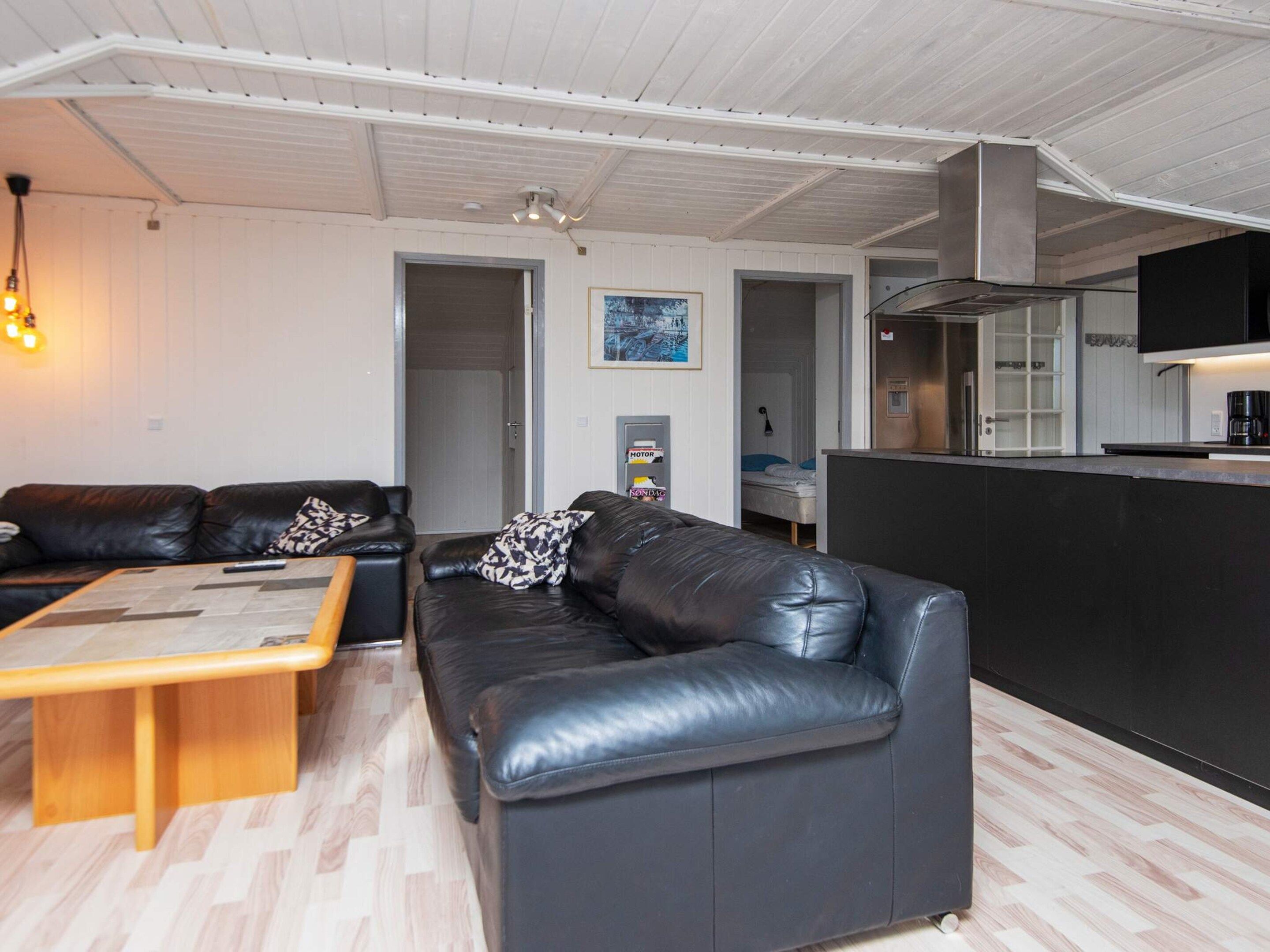 A modern living room with a black leather sofa, a wooden coffee table, and a kitchen area with a black countertop and stainless steel appliances.