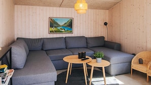 A modern living room with a grey sectional sofa, two round wooden tables, a black rug, a wicker chair, and a painting of a landscape on the wall. - 4 Star Holiday Home in Bjert (Bjert)