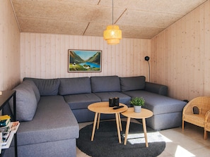 Uma sala de estar moderna com um sofá modular cinza, duas mesas redondas de madeira, um tapete preto, uma poltrona de vime e um quadro de uma paisagem na parede.