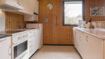 A compact kitchen with white cabinets, a stainless steel oven, a microwave, and a sink. There's a window with a view of trees, and a clock on the wall.