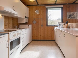 A compact kitchen with white cabinets, a stainless steel oven, a microwave, and a sink. There's a window with a view of trees, and a clock on the wall. - 6 Person Holiday Home in Toftlund (Toftlund)
