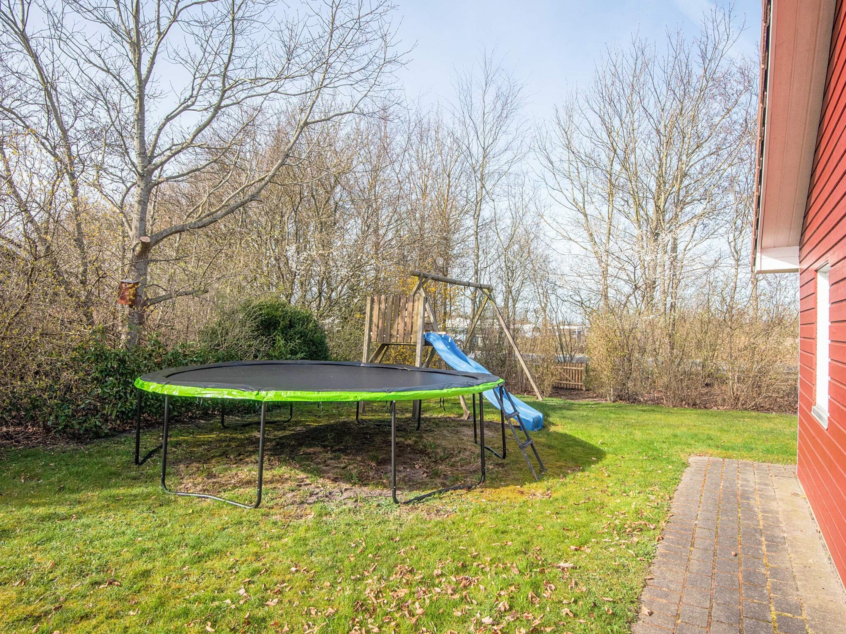 A trampoline in a backyard with a wooden swing set and a slide.