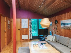A cozy room with wooden walls, a large window with a view of greenery, a sofa, a coffee table with remote controls, and a TV. - Charming Holiday Home in BlÃ¥vand with Hot Tub (Blavand)