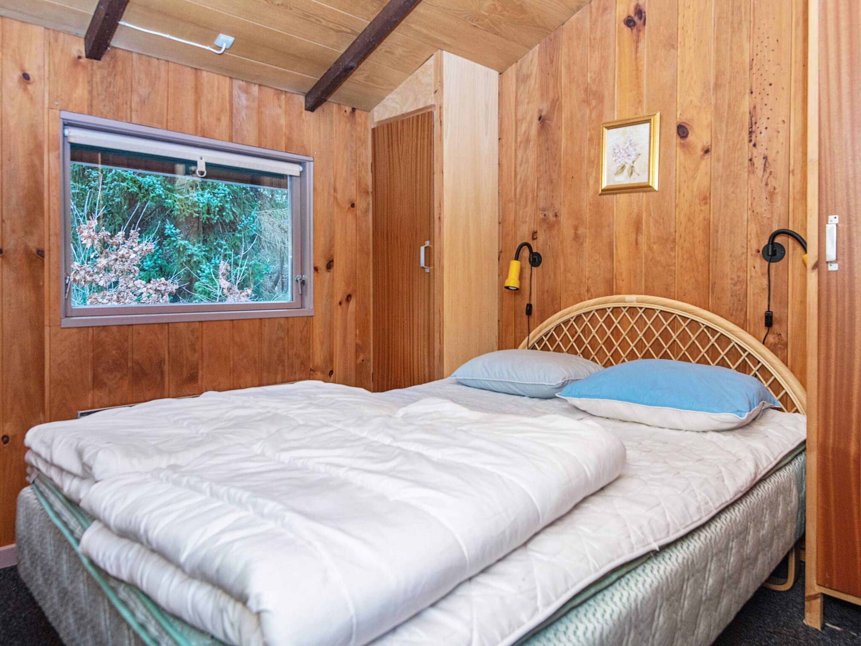 A wooden-paneled room with a bed, a window with a view of greenery, and a wall-mounted lamp.
