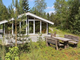 Una cabaña moderna con grandes ventanales, un patio con bancos de madera y un bosque circundante.