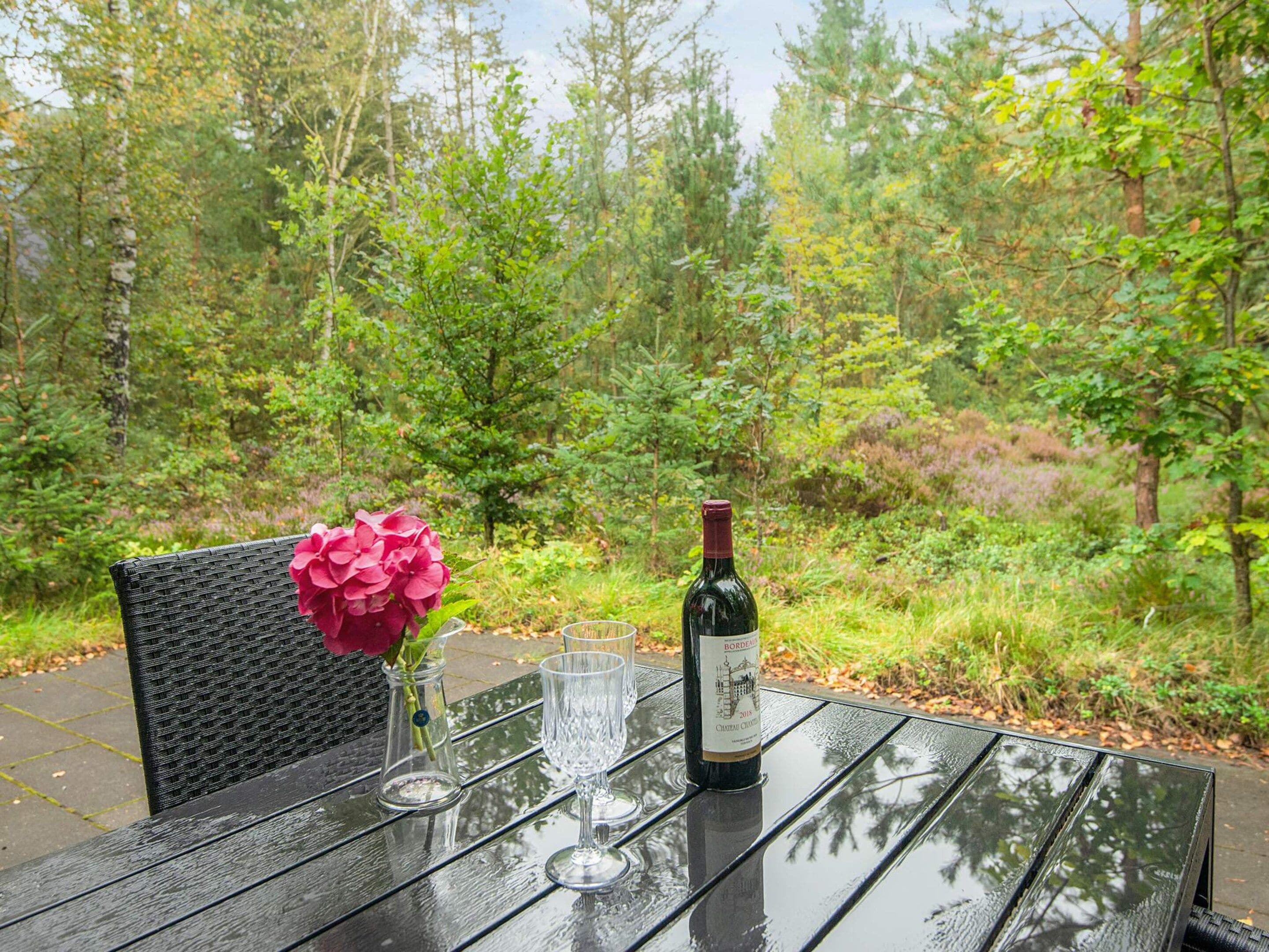 A table set outdoors with a bottle of wine, glasses, and a vase of flowers.