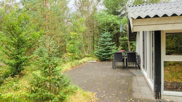 A covered outdoor seating area with a table and chairs, surrounded by dense forest.