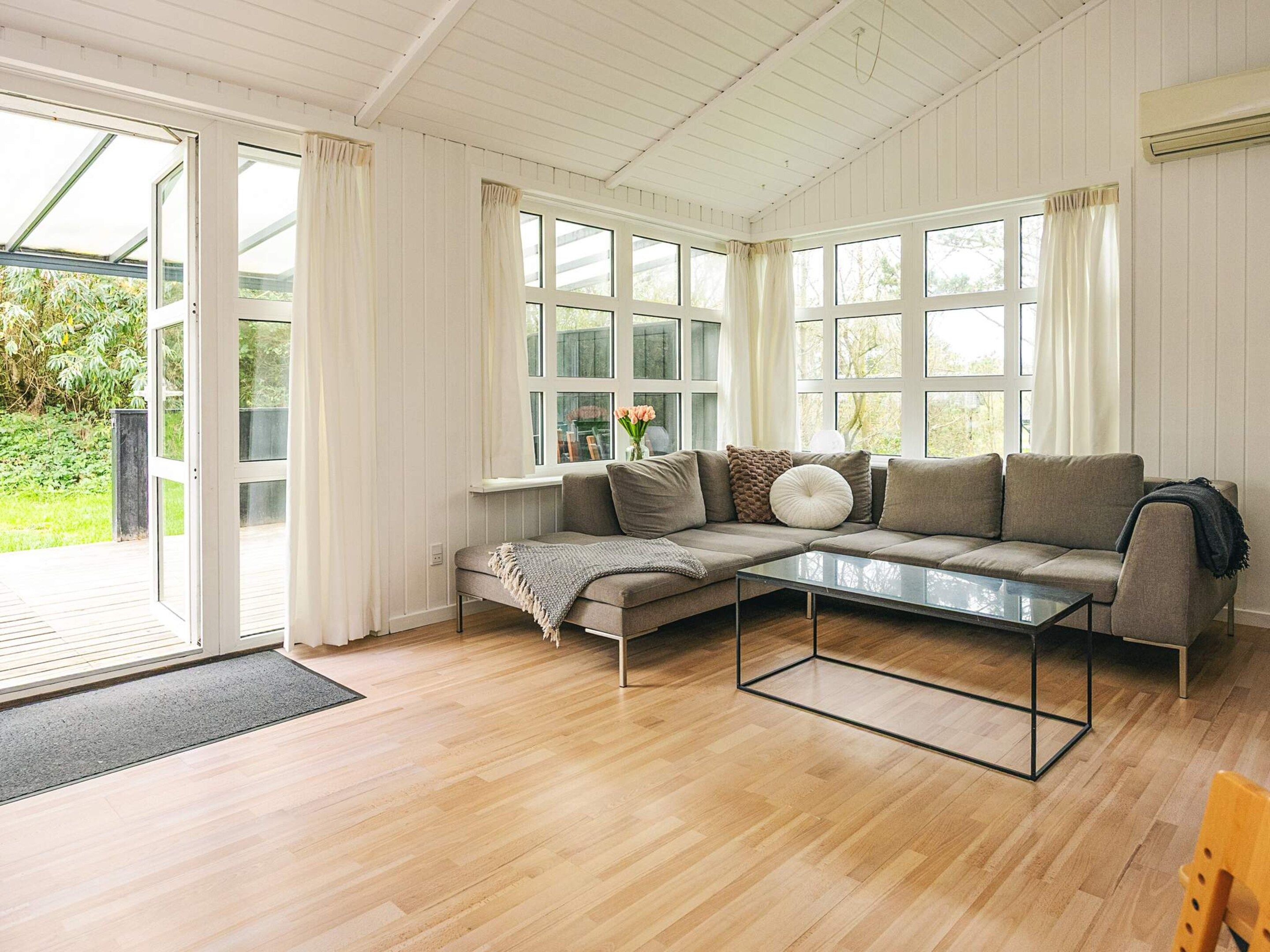 Ein modernes Wohnzimmer mit einem Ecksofa, einem Glastisch und großen Fenstern mit Vorhängen.