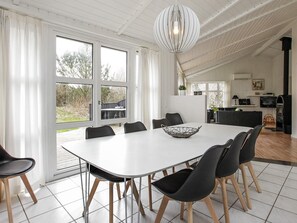 Un espace repas avec une table blanche et des chaises noires, un luminaire suspendu et une vue sur l’extérieur grâce à de grandes fenêtres.