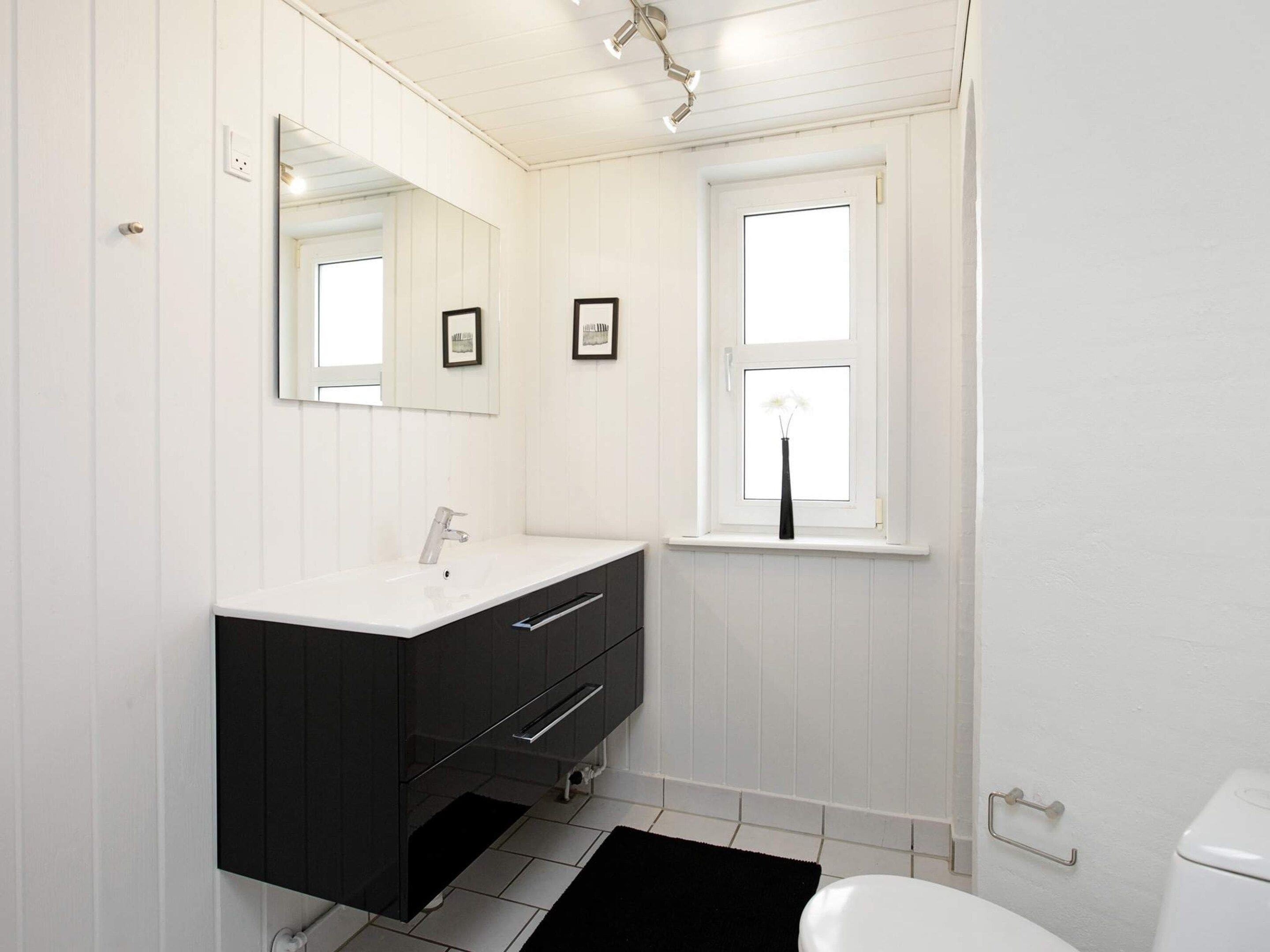 Une salle de bain avec un meuble-lavabo blanc, un miroir, une fenêtre agrémentée d’un vase et des toilettes.