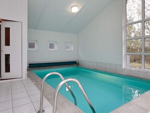 An indoor swimming pool with a tiled floor, a metal pool ladder, and a view of trees through a large window.
