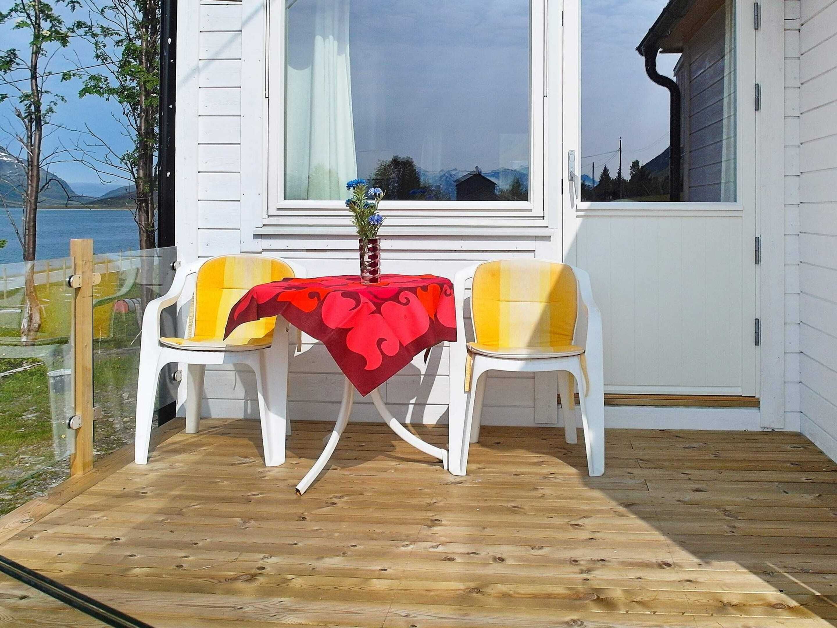A wooden deck with a table and two chairs, a window with curtains, and a view of a body of water and mountains.
