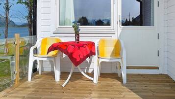 A wooden deck with a table and two chairs, a window with curtains, and a view of a body of water and mountains.