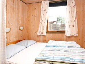 A room with wooden walls, a bed with a striped blanket, two pillows, a window with floral curtains, and a wall-mounted light fixture.