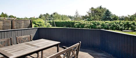A wooden deck with a table and chairs, surrounded by a dark fence and greenery.