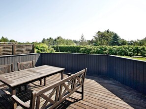 Um deck de madeira com mesa e cadeiras, cercado por uma grade escura e vegetação.