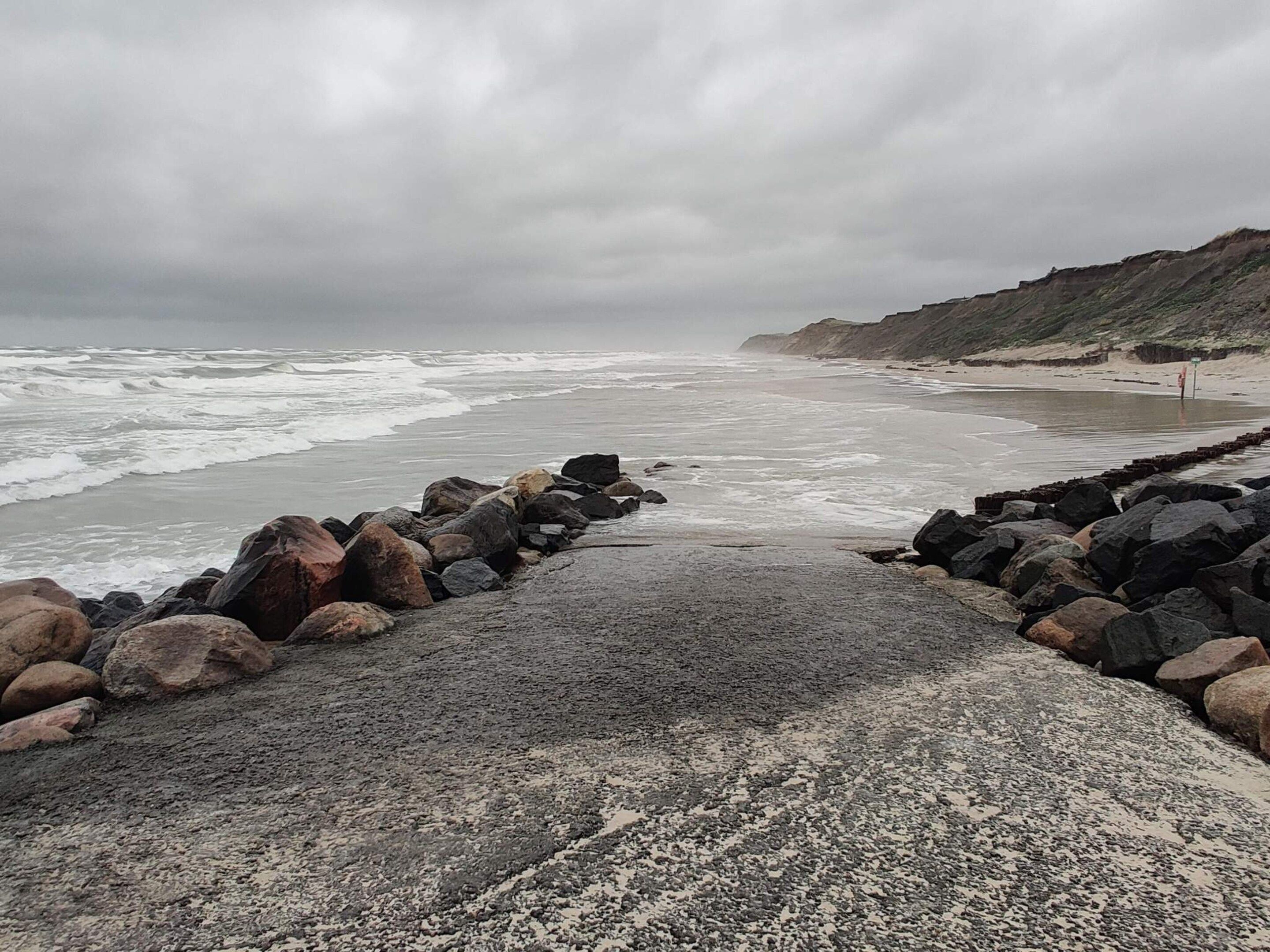 A coastal walkway with a rocky barrier, a sandy beach, and a cliff in the background.