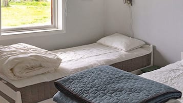 A single bed with a white mattress and a blue quilted blanket, a window with a view of greenery, and a wall-mounted light fixture.