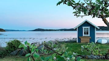 A small blue cabin with a wooden bench in front, situated by a lake with a clear view of the water and distant hills.