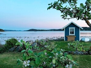 A small blue cabin with a wooden bench in front, situated by a lake with a clear view of the water and distant hills. - Holiday Home in Ljungskile (Ljungskile)