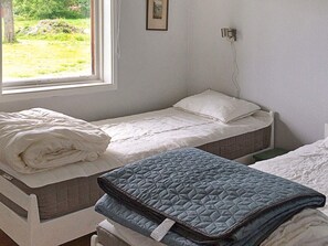 A single bed with a white mattress and a blue quilted blanket, a window with a view of greenery, and a wall-mounted light fixture.
