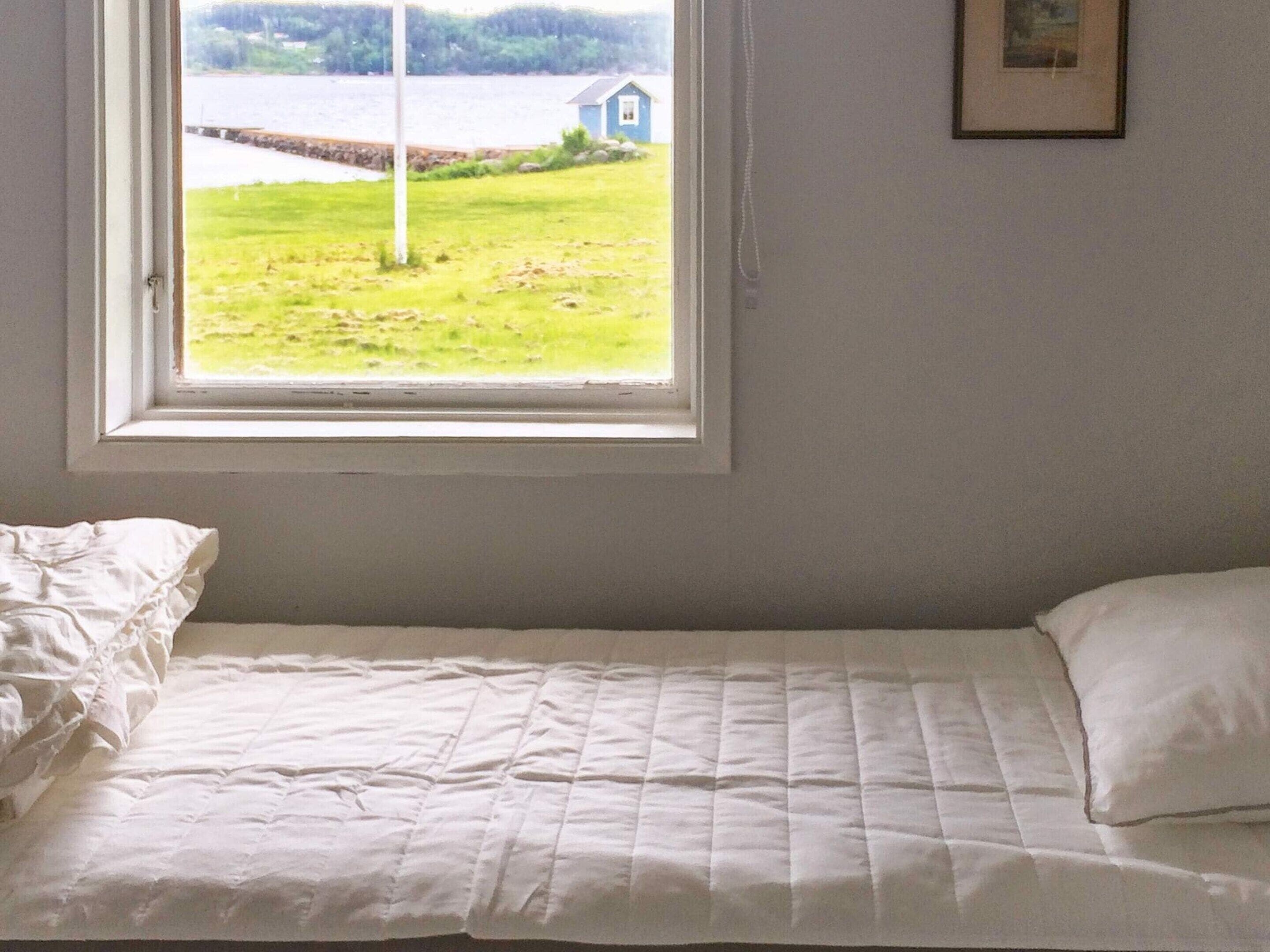 A single bed with a white quilt and pillow, a window with a view of a grassy area and a building, and a framed picture on the wall.