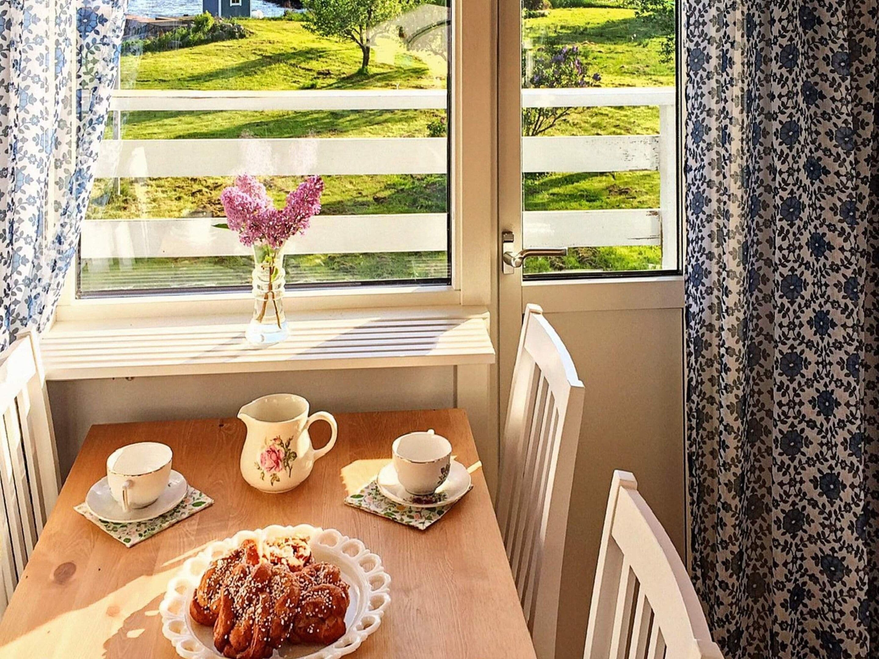 A room with a table set for tea, a vase with flowers, and a view of a green landscape through a window.
