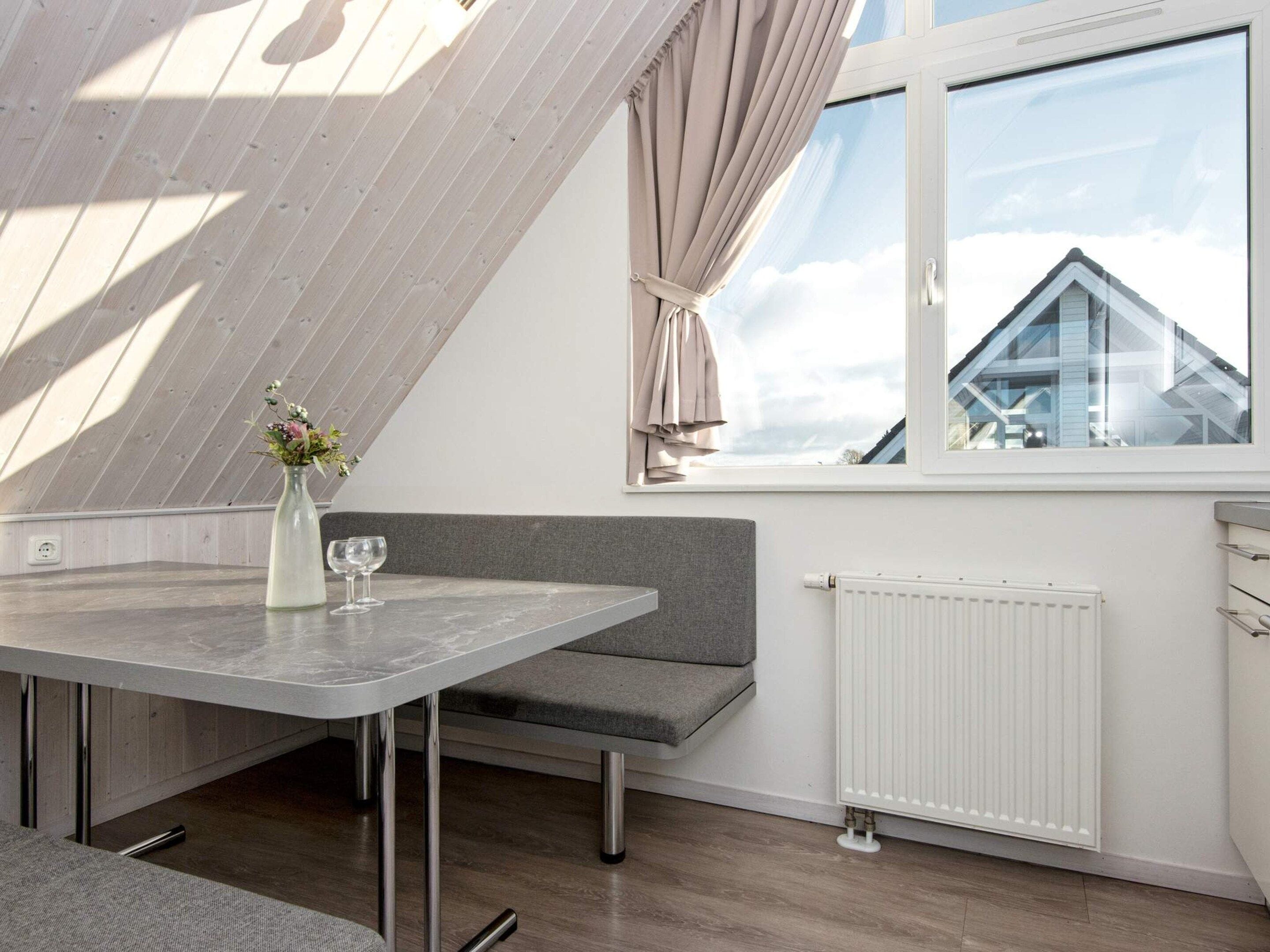 Ein modernes Zimmer mit schrägverdeckter Decke, einem Esstisch mit Stühlen, einem grauen Sofa, einem Fenster mit Vorhängen und einem Heizkörper.