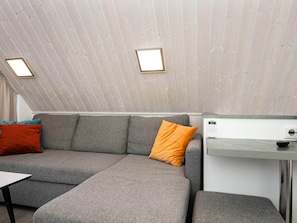 A room with a grey sofa, a small table, a white wall with a thermostat, and a ceiling with wooden panels.