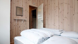 Quarto com parede de madeira, cama com roupa de cama branca, porta branca e prateleira pequena.