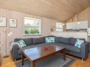 A modern living room with a grey sectional sofa, a wooden coffee table, and a kitchen area with white cabinets. - 5 Star Holiday Home in Dannemare (Dannemare)