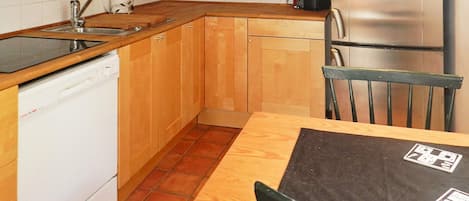 A compact kitchen with wooden cabinets, a dishwasher, a sink, a table with chairs, and a refrigerator.