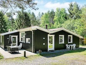 Une petite cabane aux couleurs sombres, dotée d’une terrasse carrelée, d’une table avec des chaises et d’un petit jardin.
