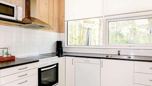 Una cocina con armarios blancos, encimera negra, microondas, estufa, lavavajillas y ventana con vistas a árboles.