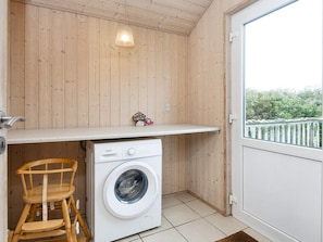A compact laundry area with a washing machine, wooden chairs, a shelf with decorative items, and a view through a door.