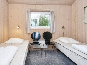 A room with two single beds, a small table, two chairs, a window with curtains, and a wall-mounted light fixture.