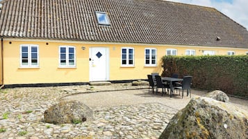 En gul bygning med et tag af fliser, en dør med et vindue, og et belagt område med sten samt et bord med stole.