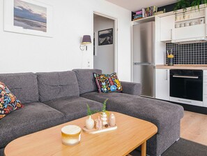 A modern living room with a grey sofa, a wooden coffee table, and a kitchen area with a fridge and oven. - 4 Star Holiday Home in Hejls (Hejls)