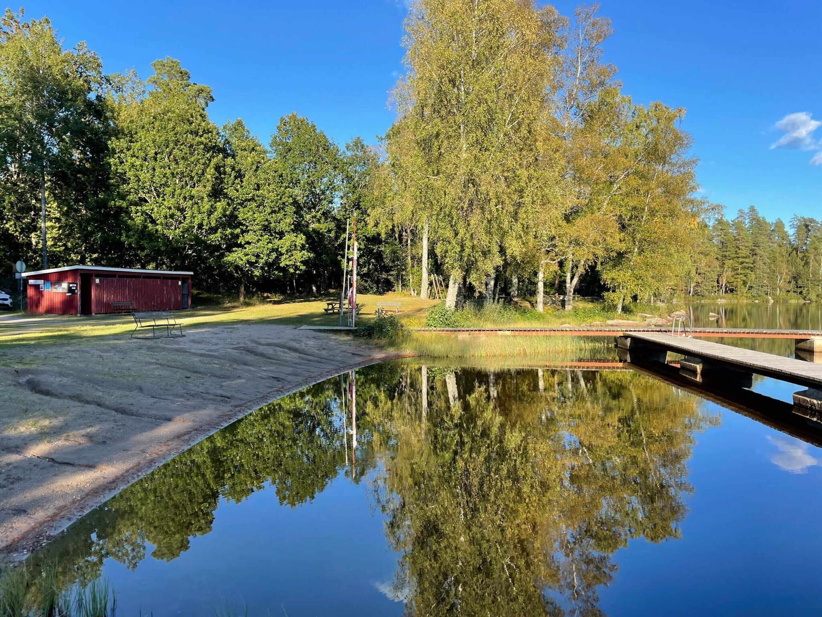 A tranquil lakeside setting with a dock, a small red building, and dense trees reflecting in the calm water.