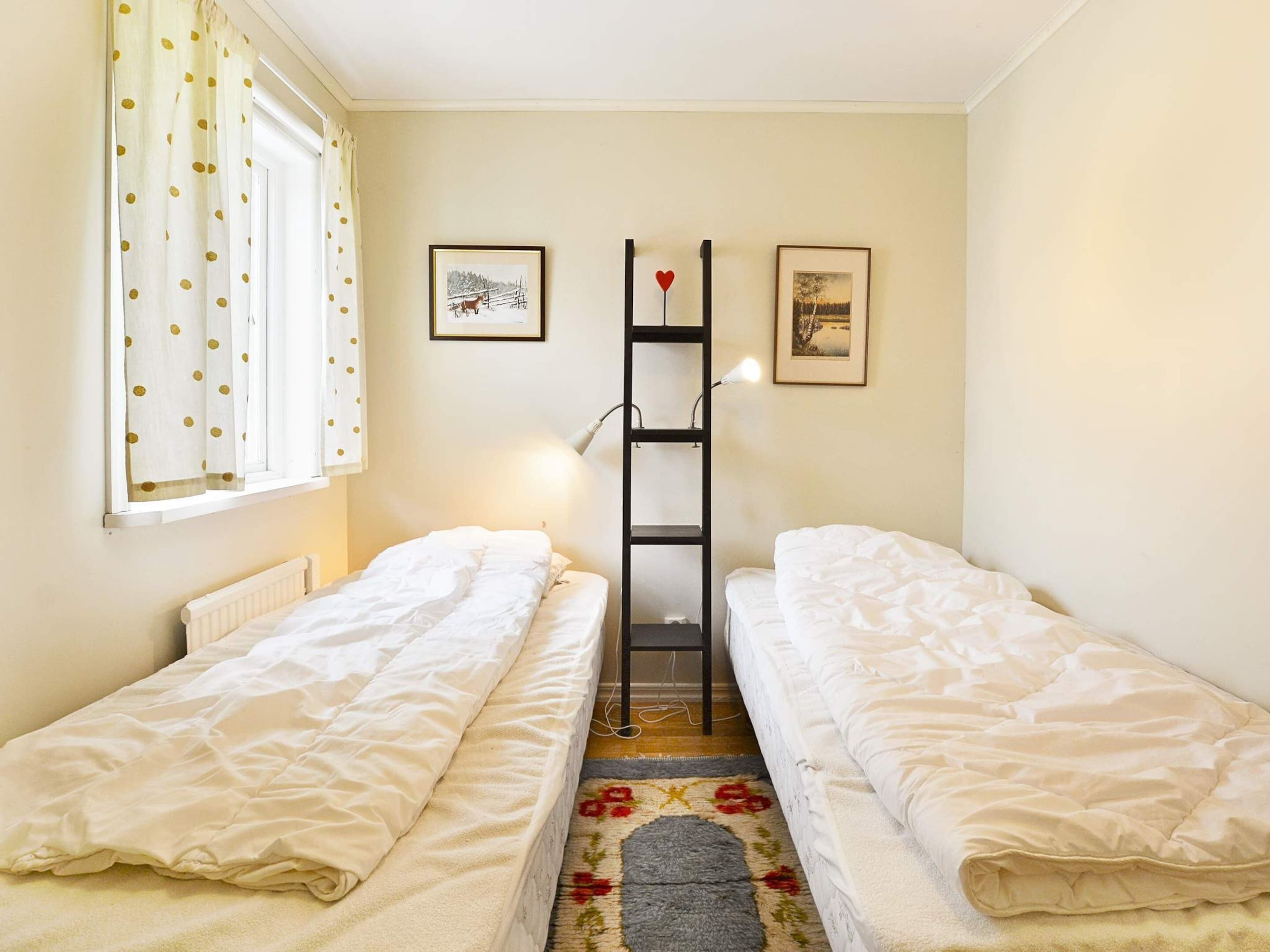 Una habitación con dos camas individuales, un radiador, una ventana con cortinas, una lámpara y dos cuadros enmarcados en la pared.