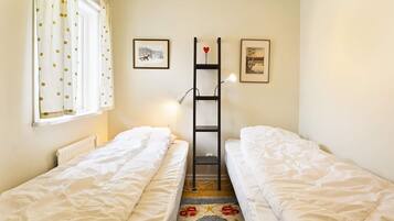 Una habitación con dos camas individuales, un radiador, una ventana con cortinas, una lámpara y dos cuadros enmarcados en la pared.