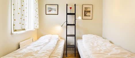 A room with two single beds, a radiator, a window with curtains, and a shelf with pictures.