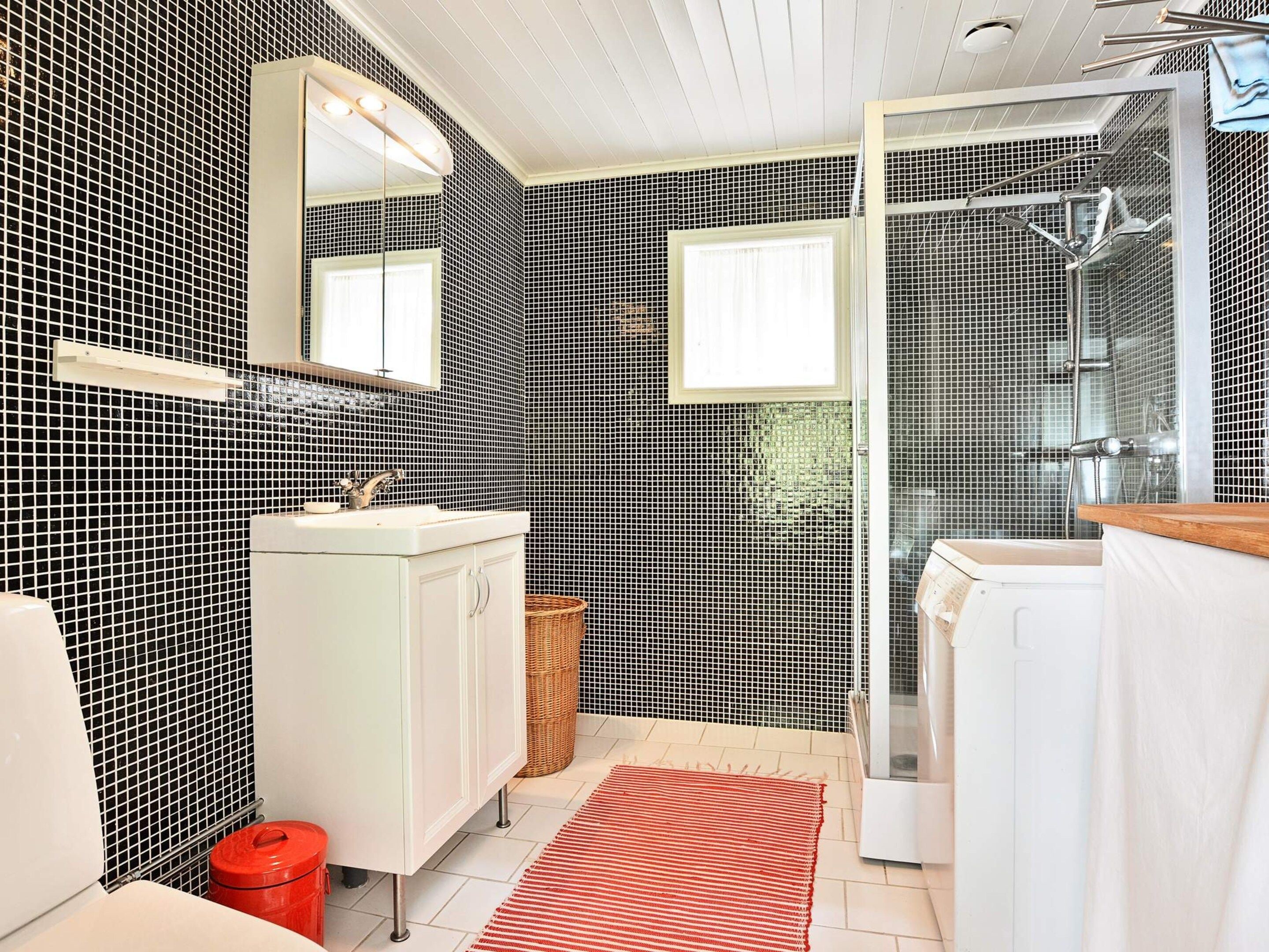A bathroom with black and white tiled walls, a white vanity with a mirror, a red rug, and a shower area.