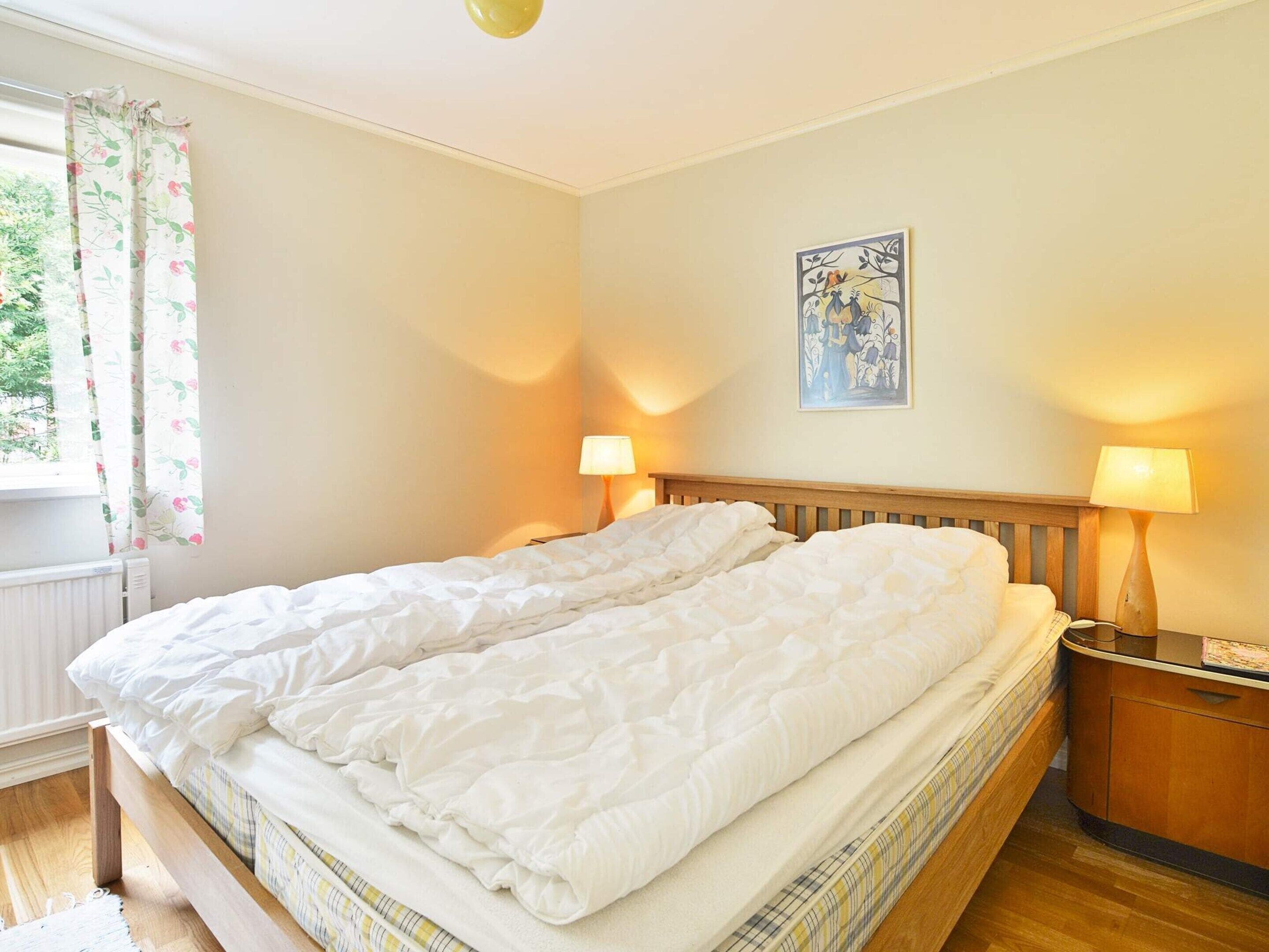 Cama doble con cabecera de madera, dos lámparas de noche, un radiador y una ventana con cortinas florales.