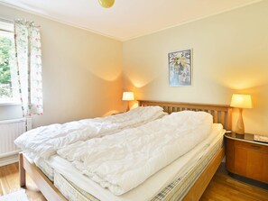 A double bed with a wooden headboard, two bedside lamps, a radiator, and a window with floral curtains. - 4 Person Holiday Home in Hallabro (Hallaröd)