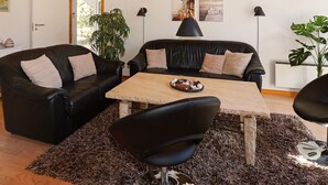 A modern living room with a wooden ceiling, two black leather sofas, a wooden coffee table, and a black armchair. There are two black pendant lights, a large potted plant, and a framed picture on the wall. - Modern Holiday Home in Jutland With Sauna (Hadsund)