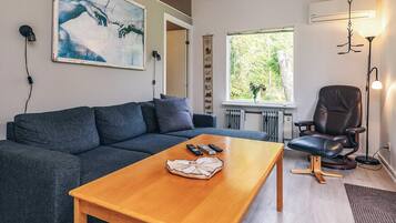 A modern living room with a grey sofa, a wooden coffee table, a black leather chair, and a large window with a view of greenery.